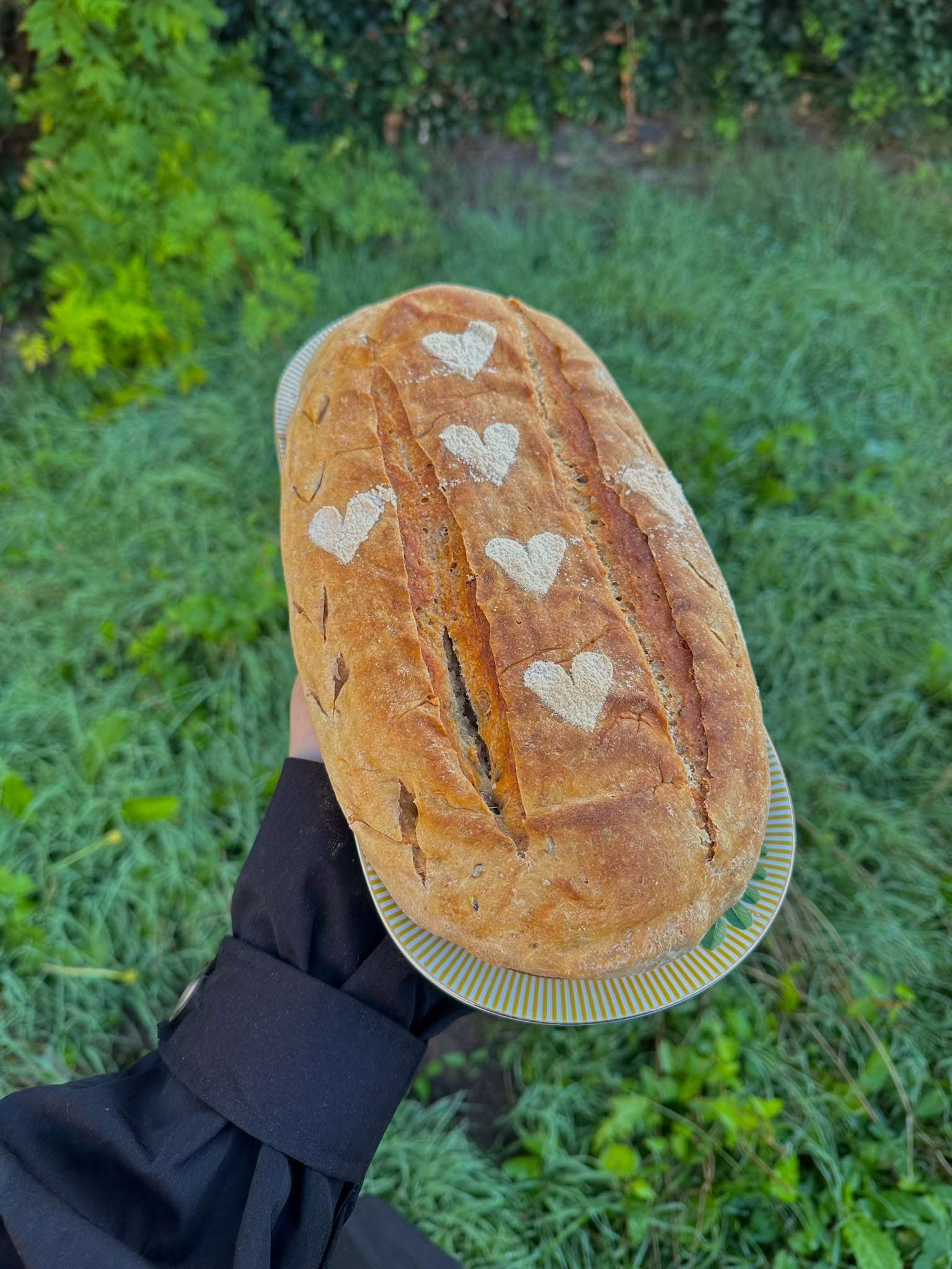 Een lichtbruin zuurdesembrood met lange insnijdingen en bloemhartjes, op een gestreept bord in de tuin